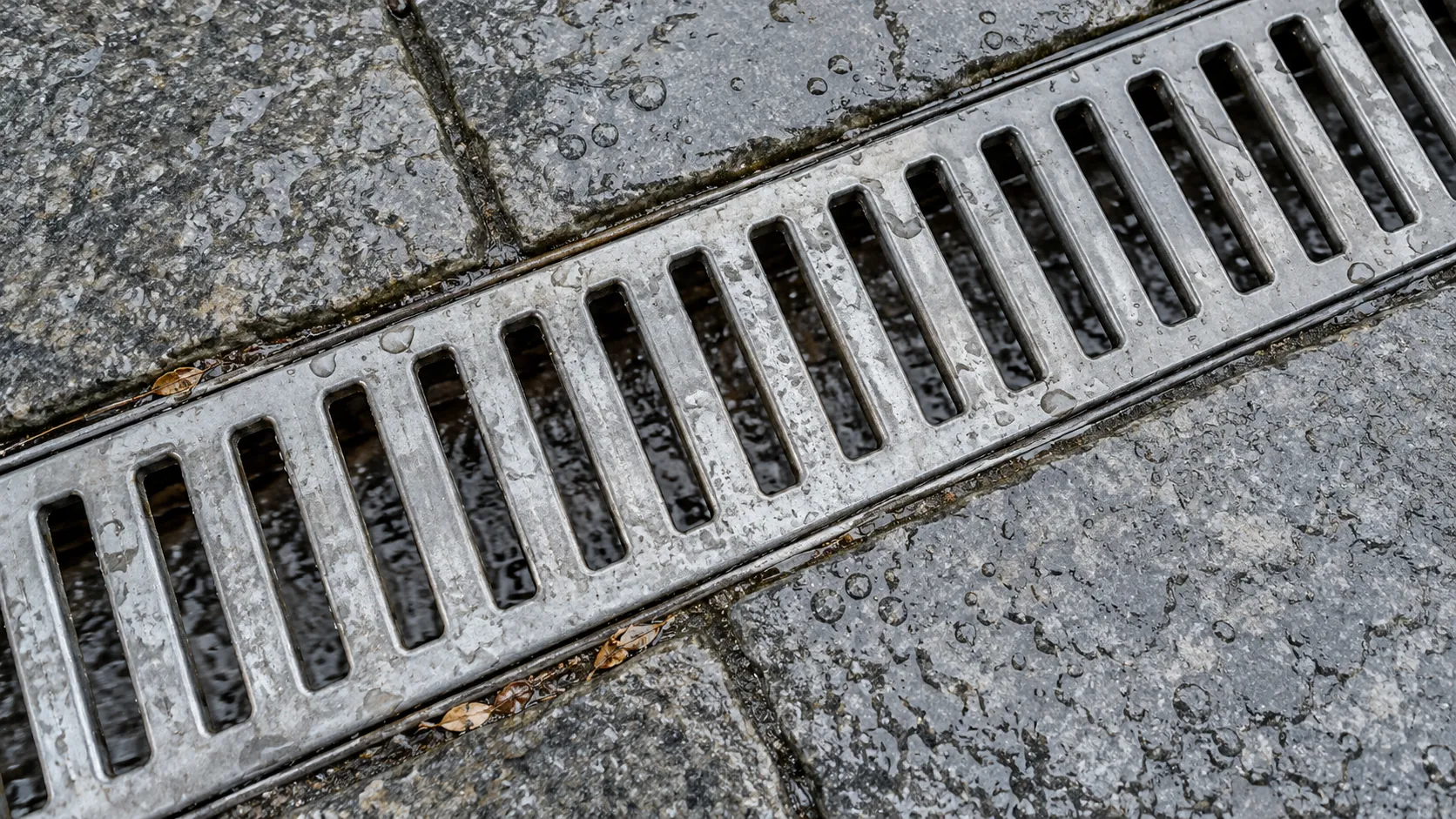 A galvanized outdoor drain grate set between damp gray paving slabs